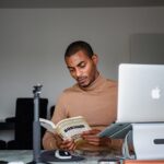 Man reading a book at a desk with a laptop in an indoor setting.