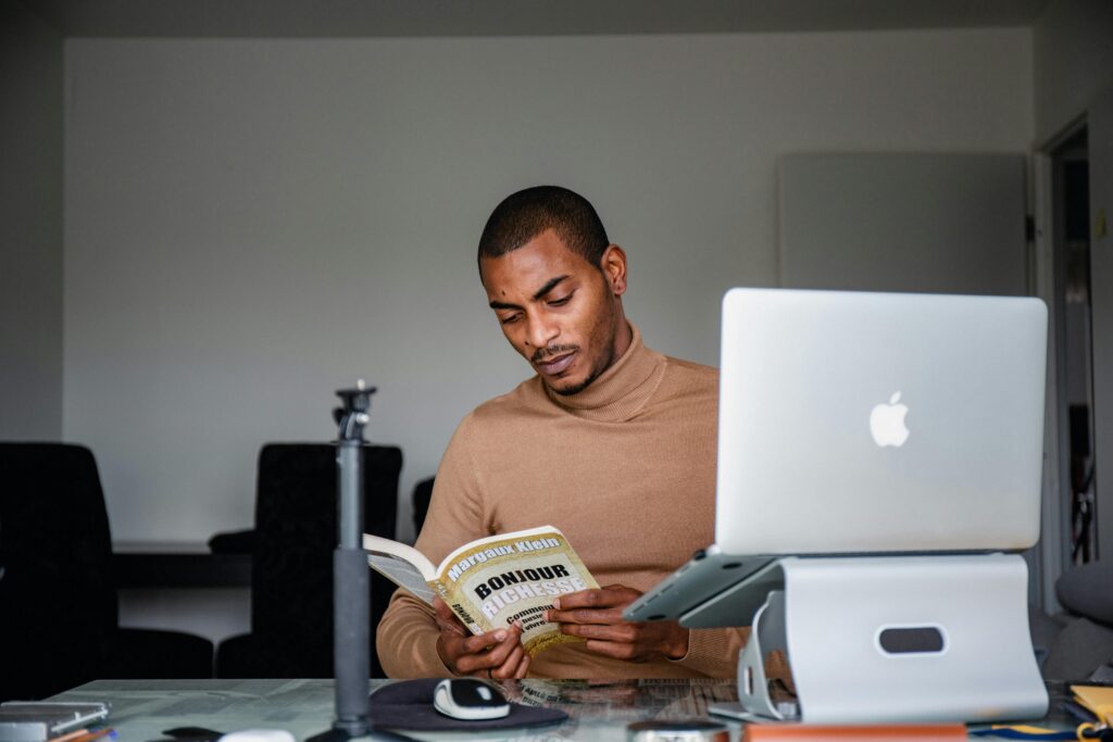 Man reading a book at a desk with a laptop in an indoor setting.