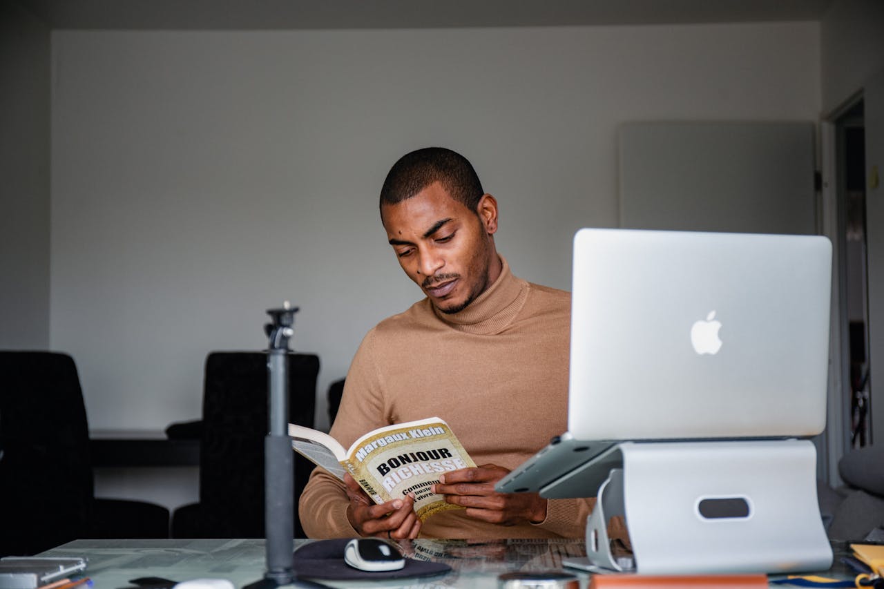 Man reading a book at a desk with a laptop in an indoor setting.