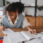 Focused young woman taking notes from a book at home, surrounded by study materials.